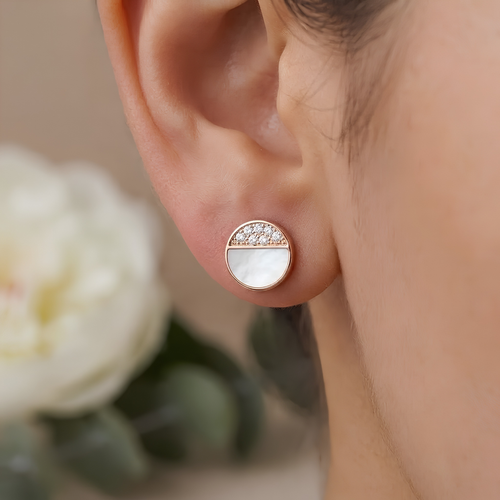 A close-up of a person's ear wearing a small round stud earring with a split design: one half is white mother-of-pearl and the other half is rose gold with tiny clear stones.