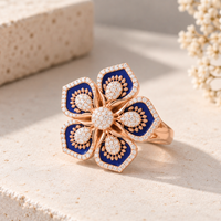 A five-petal flower ring in rose gold with deep blue enamel and pavé-set clear stones, displayed on a textured beige stone surface with soft-focus dried flowers in the background.