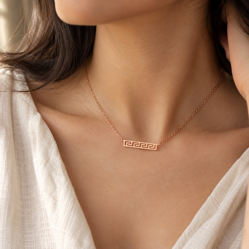 A close-up lifestyle shot of a woman wearing a delicate rose gold necklace with a rectangular Greek key pendant, styled with a white linen top.