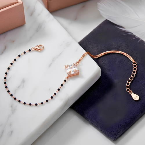A delicate rose gold chain bracelet with a central square clear crystal and small black beads along the chain, styled on a white marble block next to a dark navy velvet pouch.