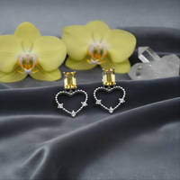 Heart-shaped earrings with yellow gemstones on a dark fabric background with flowers and crystals.