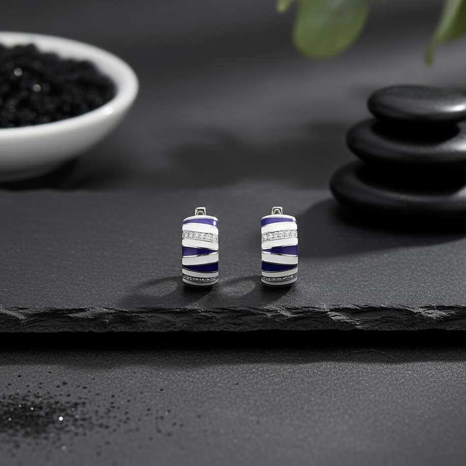 Pair of blue and silver cylindrical earrings on a dark surface with stones and a bowl in the background.