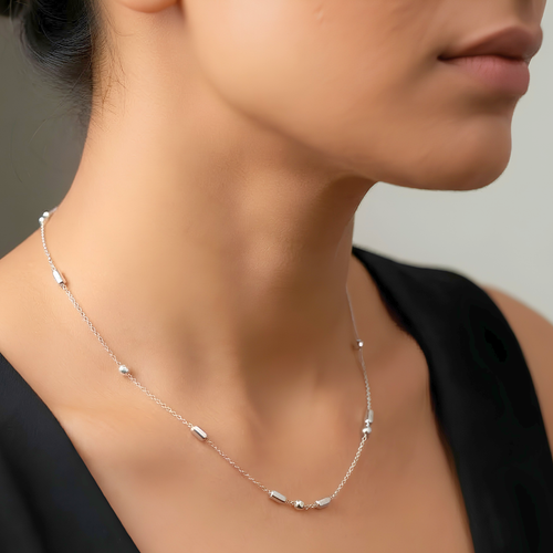 A close-up of a model's neck and lower face wearing a delicate silver chain necklace with small cylindrical bead accents.