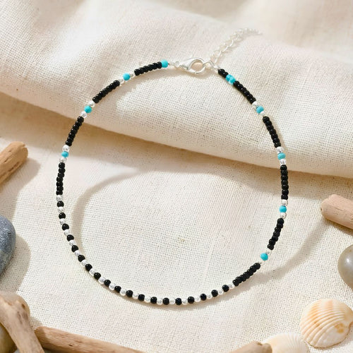 Beaded bracelet with black and turquoise beads on a textured beige surface with seashells and driftwood.