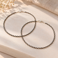 Two twisted rope-style hoop anklets in a silver-tone finish, arranged overlapping on a light beige stone surface with soft natural shadows and dried flowers in the corner.